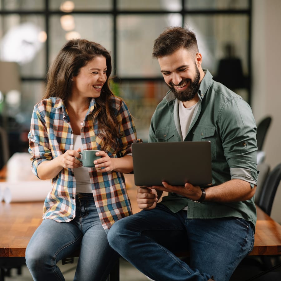 An adult man and woman are leaned up against a table. She is laughing and holding a mug, and he is smiling and looking at a laptop.