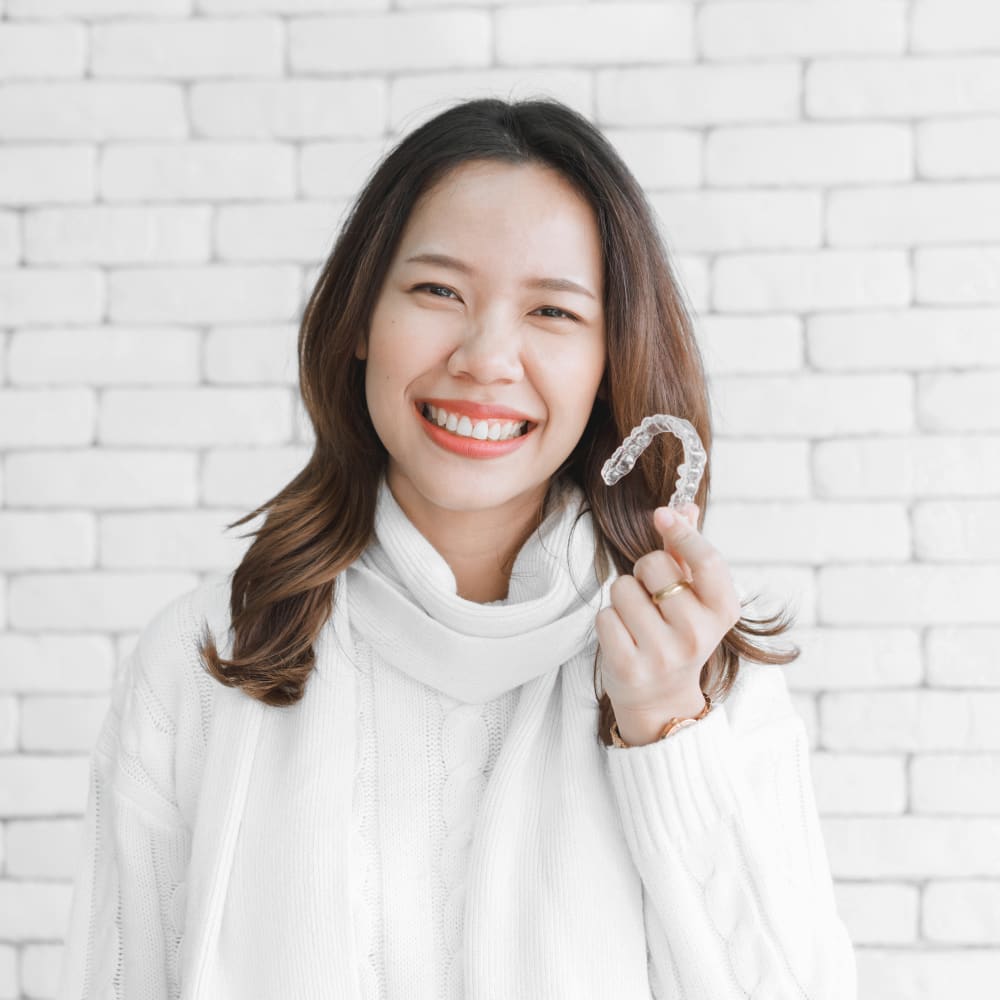 A woman wearing a white sweater is smiling and standing against a white brick wall. She is holding a tray of clear aligners.