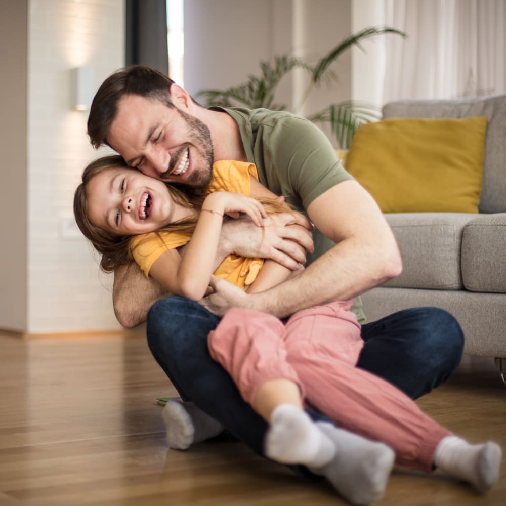 A man is sitting on the floor hugging his young child. They are both smiling and laughing.