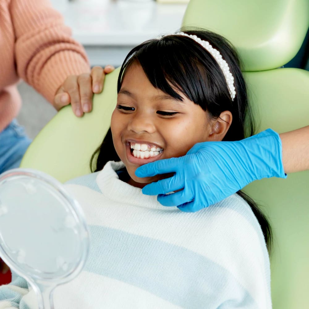 A young girl is in a dentist chair looking into a mirror as she shows off her teeth.