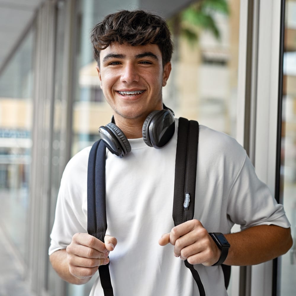 A teenage boy with braces is holding his backpack straps and smiling. Around his neck hang a pair of large headphones.