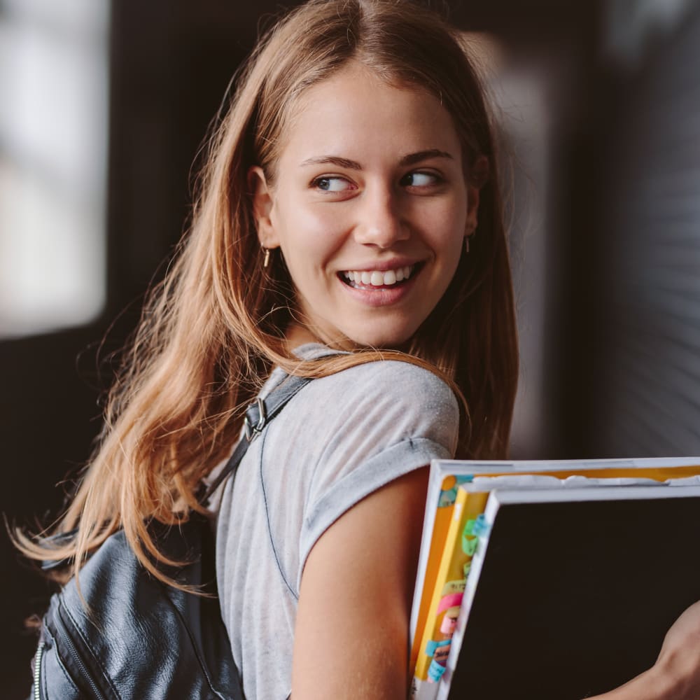 A teenage girl wearing a backpack and carrying books looks over her shoulder and smiles.